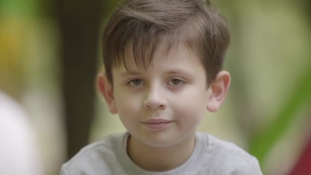Portré aranyos mosolygó fiú nézte a kamerát ül a szabadban. Gondtalan gyermekkor. Egy gyerek pózol a kamera — Stock videók