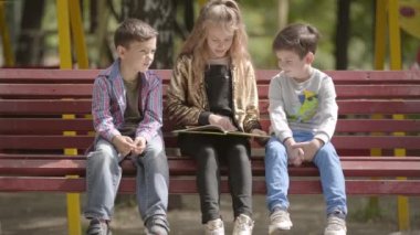 Sevimli genç kız bir bankta açık havada otururken iki küçük kardeşler için bir kitap okuyor. Parkta güneşli bir yaz gününde çocuklar boş zaman.