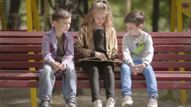 Das süße Teenie-Mädchen liest zwei jüngeren Brüdern ein Buch vor, während sie draußen auf einer Bank sitzt. Freizeit für Kinder an einem sonnigen Sommertag im Park. — Stockvideo