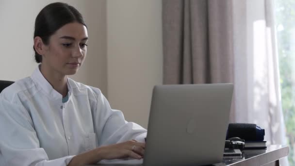 Médica profissional bem sucedida digitando em um laptop enquanto se senta em uma mesa em seu escritório. Uma jovem está distraída do trabalho e olhando para a câmera sorrindo. Conceito de profissão, medicina — Vídeo de Stock