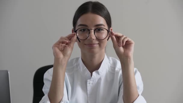 彼女の手に眼鏡を持つかわいい若いプロの女性医師の肖像画は、カメラを見ます。フォーカスは顔から眼鏡に移動します。職業、医療、ヘルスケアの概念. — ストック動画