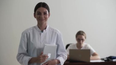 Kameraya bakarken dizüstü bilgisayar tutan genç profesyonel kadın doktor portresi. Arka planda gözlüklü bir kız dizüstü bilgisayarda yazıyor. Meslek kavramı, tıp, bakım.