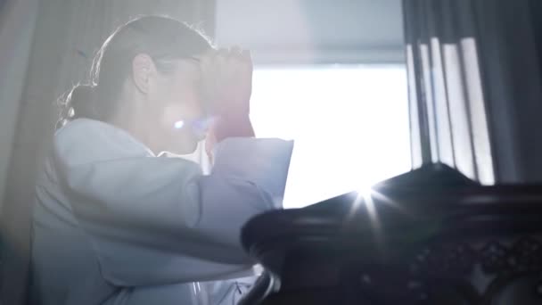 Professionele vrouwelijke arts in glazen typen op een laptop terwijl zittend aan een tafel tegen de achtergrond van een venster en zonlicht in zijn kantoor. Concept van beroep, geneeskunde en gezondheidszorg. — Stockvideo