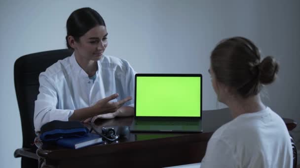 Profesional médico femenino cuenta algo a la paciente femenina y que muestra con la mano en la pantalla verde del portátil. Concepto de profesión, medicina, salud . — Vídeos de Stock