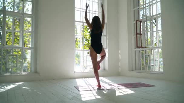 Mujer caucásica sana de pie sobre la esterilla en la gran sala blanca practicando yoga. Mujer joven haciendo ejercicio en el gimnasio en el interior. Una joven está haciendo ejercicio en casa. Estilo de vida saludable — Vídeo de stock