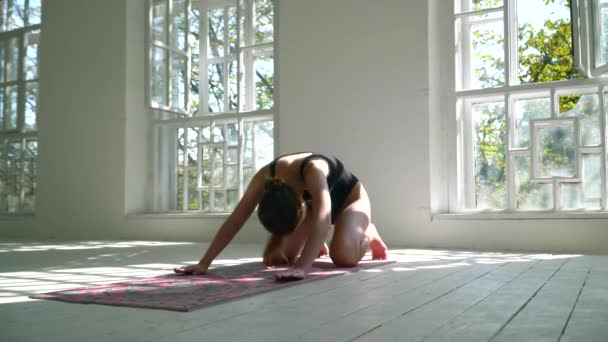 Mujer caucásica sana practicando yoga en la esterilla en la habitación grande blanca. Mujer joven en ropa deportiva haciendo ejercicio en el gimnasio en el interior . — Vídeos de Stock