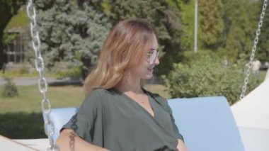 Mutlu gülümseyen kız parkta veya bahçede salıncak üzerinde oturan, onun zaman zevk. Güneş gözlüğü takan genç kadın hafta sonunu dışarıda geçiriyor. Pozitif güneşli bir gün. Izin günü. Tatil konsepti