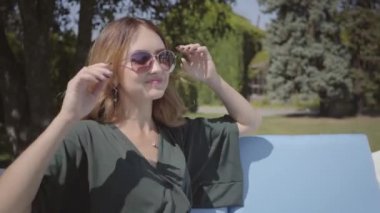 Güneş gözlüğü takan genç kadın hafta sonunu dışarıda geçiriyor. Mutlu sevimli genç kız parkta veya bahçede salıncak üzerinde oturan, onun zaman zevk. Güneşli bir gün. Izin günü. Tatil konsepti. Gerçek insanlar serisi.