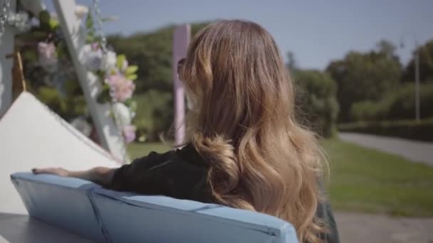 Menina bonita sentada no balanço no parque ou jardim, desfrutando de seu tempo. Jovem mulher passar o fim de semana ao ar livre. Dia ensolarado positivo. Dia de folga. Conceito de férias. Câmara a aproximar-se — Vídeo de Stock