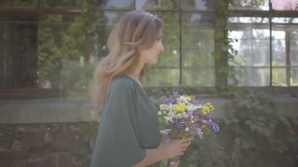 建物の近くに野生の花の花束を持って歩く美しい若い女性の肖像画。夏の概念、休息日. — ストック動画