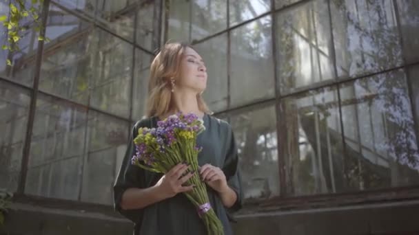 古い建物に立って野生の花を嗅ぐかわいい若い女性の肖像画。夏の概念、休息日 — ストック動画