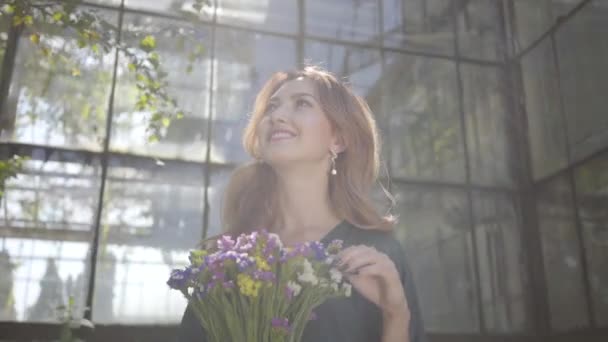 古い建物に立っている野生の花の花束を持つかわいい若い女性の肖像画。夏の概念、女性らしさ、休息日。リアルピープルシリーズ. — ストック動画