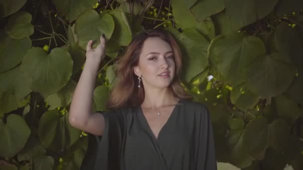 Portré csinos fiatal nő nézi a kamerát, és mosolyogva állva a fán nagy levelek. A nyár fogalma, nőiesség, pihenőnap. Valódi emberek sorozata. — Stock videók