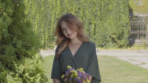 Ritratto di giocosa giovane donna carina che tiene mazzo di fiori selvatici guardando la macchina fotografica mentre in piedi all'aperto. Concetto di ora legale, giorno di riposo. Serie persone reali . — Video Stock