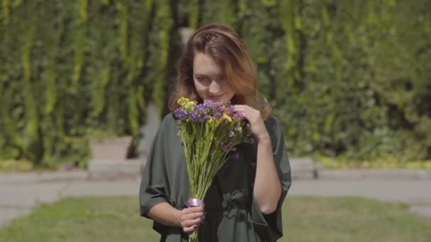 Retrato de chica linda juguetona oliendo flores silvestres mirando a la cámara mientras está de pie al aire libre. Concepto de verano, día de descanso. Serie de personas reales . — Vídeos de Stock