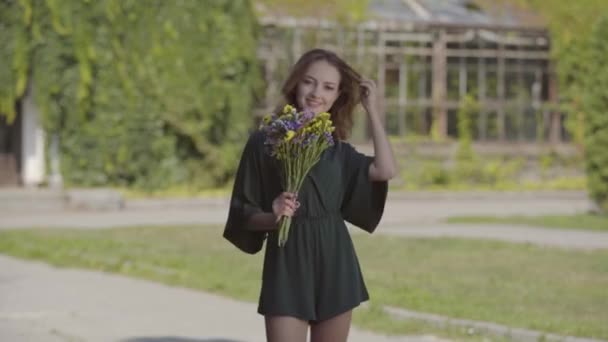 Πορτρέτο παιχνιδιάρικο νεαρή γυναίκα κρατώντας μπουκέτο από άγρια λουλούδια κοιτάζοντας την κάμερα, ενώ στέκεται σε εξωτερικούς χώρους. Η έννοια του καλοκαιριού, η μέρα ανάπαυσης. Σειρές πραγματικών ατόμων. — Αρχείο Βίντεο
