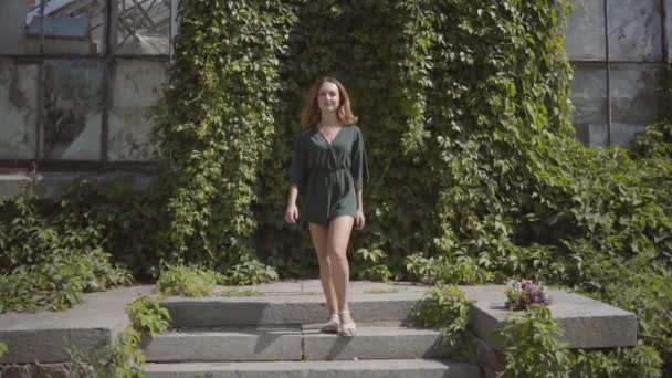 Уверенная красивая молодая женщина идет к камере улыбаясь, а затем подмигивает в зеленом летнем парке. Концепция летнего времени, женственности, отдыха. Медленное движение . — стоковое видео