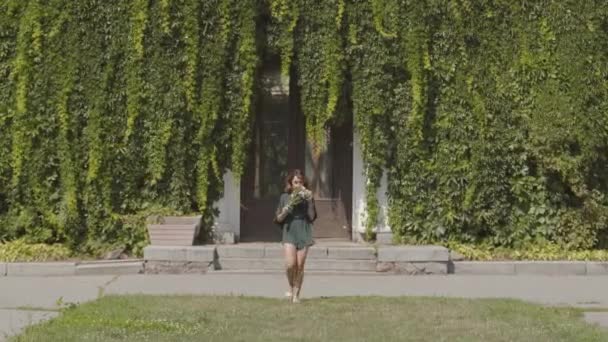 Mujer joven y segura de sí misma caminando hacia la cámara sonriendo sosteniendo un ramo de flores silvestres en el verde parque de verano. Concepto de verano, feminidad — Vídeo de stock