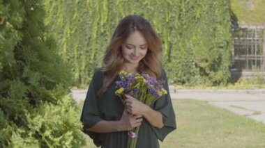 Açık havada dururken kameraya bakarak yabani çiçek bukettutan oldukça smilling genç kadın portresi. Yaz saati kavramı, dinlenme günü. Gerçek insanlar serisi.