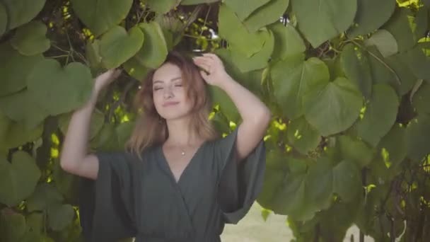 Retrato de una hermosa mujer joven mirando a la cámara y sonriendo mientras está de pie en el árbol con hojas grandes. Concepto de verano, feminidad, día de descanso. Serie de personas reales . — Vídeos de Stock