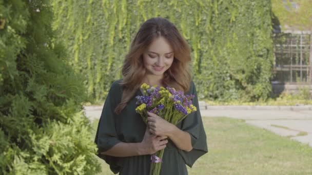 屋外に立ちながらカメラを見つめる野生の花の花束を持つかわいい笑顔の若い女性の肖像画。夏の概念、休息日。リアルピープルシリーズ. — ストック動画