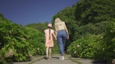 Sevimli sarışın anne el ele yaz bahçesinde küçük sevimli kızı ile yürüyüş geri görünümü. Mutlu bir aile. Kadın ve kız birlikte açık havada.