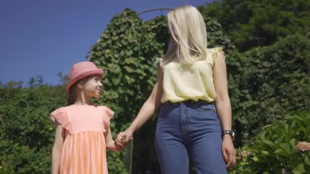 Linda madre rubia caminando con la pequeña hija adorable en el jardín de verano tomados de la mano. Familia feliz. Mujer y chica juntas al aire libre . — Vídeo de stock