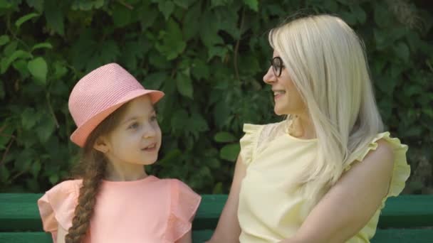 Aranyos Érett szőke anya a szemüveg és az ő kislánya beszélgető ülve a padon a nyári parkban. Boldog szerető család. Nő és lány együtt a szabadban. — Stock videók