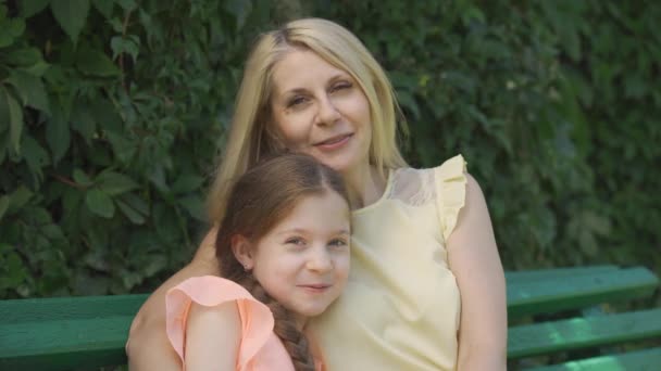 Portret van mooie rijpe blonde moeder in glazen en haar kleine dochter kijkend naar camera terwijl zittend op de Bank in het zomerpark en knuffelen. Gelukkige familie. Vrouw en meisje samen buitenshuis. — Stockvideo