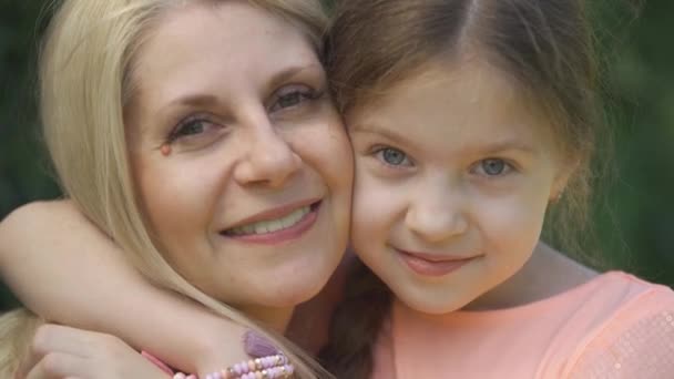 Close-up portret glimlachend blond moeder en haar kleine dochter knuffelen in het zomerpark kijkend naar camera. Gelukkig liefdevolle familie. Vrouw en meisje samen buitenshuis. — Stockvideo