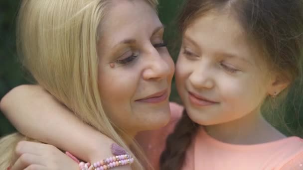 Sarışın anne ve küçük kızının yaz parkında otururken sarılıp öpüştüğü yakın çekim portresi. Mutlu sevgi dolu bir aile. Kadın ve kız birlikte açık havada. — Stok video
