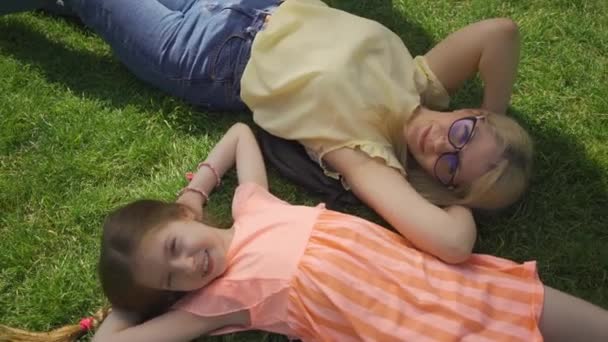 Bonita madre rubia y su adorable hija tumbada en la hierba en el jardín o parque de verano. Familia feliz. Mujer y chica juntas al aire libre . — Vídeos de Stock