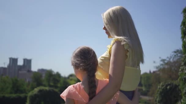 Ξανθιά μητέρα και η μικρή κόρη της να περνούν το χρόνο τους στον καλοκαιρινό κήπο ή πάρκο. Το κορίτσι που αγκαλιάζει τη μαμά της. Ευτυχισμένη οικογένεια. Γυναίκα και κορίτσι μαζί σε εξωτερικούς χώρους. — Αρχείο Βίντεο
