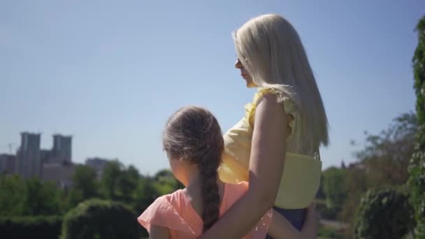 Madre bionda e la sua piccola figlia trascorrono del tempo nel giardino o nel parco estivo. La ragazza che abbraccia sua madre. Una famiglia felice. Donna e ragazza insieme all'aperto . — Video Stock