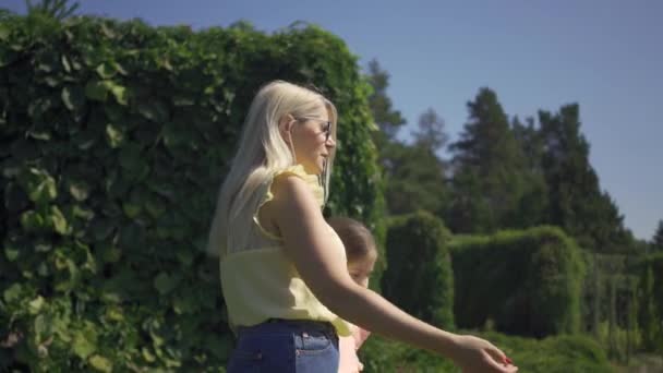 Fritid av söt mogen blond mor och hennes lilla dotter i sommarträdgården eller parken. Flickan kramar hennes mamma. Lycklig familj. Kvinna och flicka tillsammans utomhus. — Stockvideo