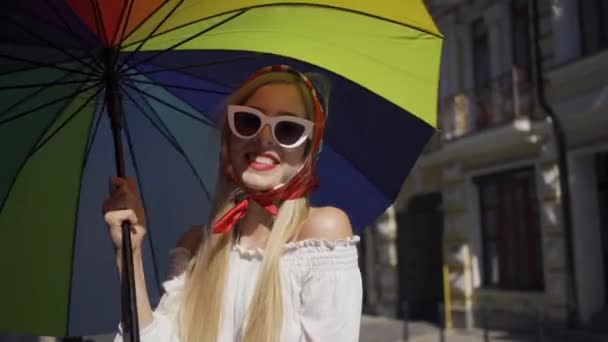 Cute young woman looking at the camera holding multicolored umbrella standing on the street. Attractive fashionable girl enjoying sunny day in the old European city. Tourism concept. — Stock Video