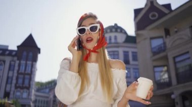 Portre moda güzel genç kadın kahve fincan ı ile güneşte sokakta duran ve cep telefonu ile konuşuyor. Eski Avrupa şehrinde güneşli bir günün tadını çıkaran çekici kız. Turizm konsepti