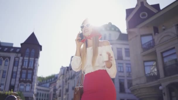 Moda bella giovane donna con una tazza di caffè in piedi sulla strada al sole e parlando al cellulare. Ragazza attraente godendo giornata di sole nella vecchia città europea. Concetto turistico — Video Stock