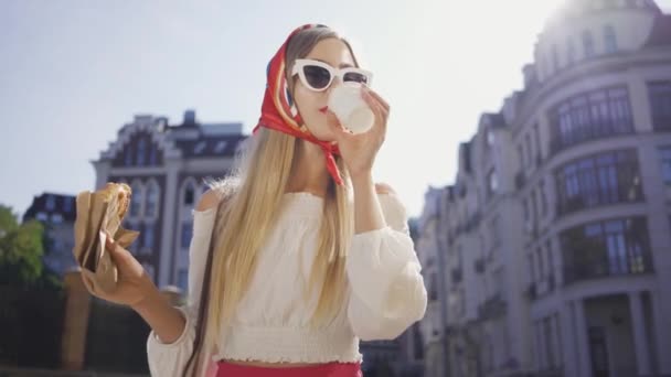 Retrato bela jovem andando na rua bebendo café e comendo croissant. Menina na moda atraente desfrutando de dia ensolarado na antiga cidade europeia. Conceito de turismo — Vídeo de Stock