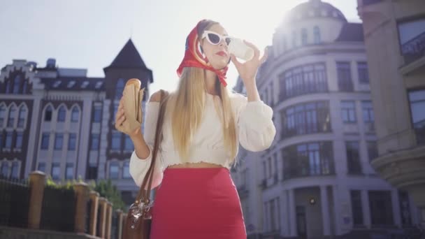 Πορτραίτο χαριτωμένο νεαρή γυναίκα που περπατάει στο δρόμο πίνοντας καφέ και τρώγοντας κρουασάν. Ελκυστικό μοντέρνο κορίτσι απολαμβάνοντας ηλιόλουστη μέρα στην παλιά Ευρωπαϊκή πόλη. Τουριστική έννοια — Αρχείο Βίντεο