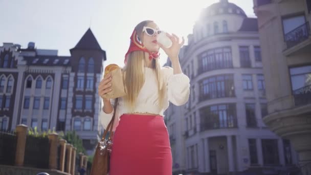 Retrato bonita jovem andando na rua beber café e comer croissant. Menina na moda atraente desfrutando de dia ensolarado na antiga cidade europeia. Conceito de turismo — Vídeo de Stock