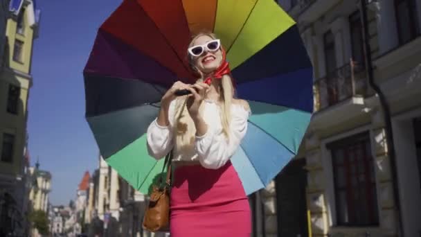 Mulher bonita positiva olhando para a câmera segurando guarda-chuva multicolorido na rua. Menina na moda atraente desfrutando de dia ensolarado na antiga cidade europeia. Conceito de turismo — Vídeo de Stock