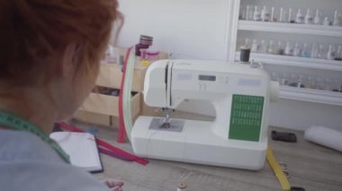 Kızıl saçlı kadın ışık odasında masada oturan giysileri diker. Terzi evde çalışıyor. Hobi. Dikiş kıyafetleri. Omzundan ateş etme.