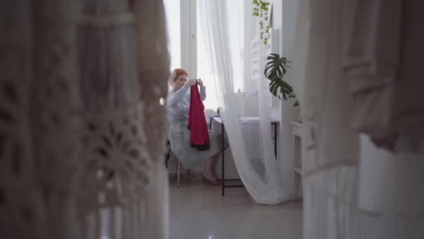 Diligente mujer pelirroja cose en la mesa en el taller. La costurera trabaja diligente y cuidadosamente. Hobby y trabajo. Costura de ropa . — Vídeo de stock