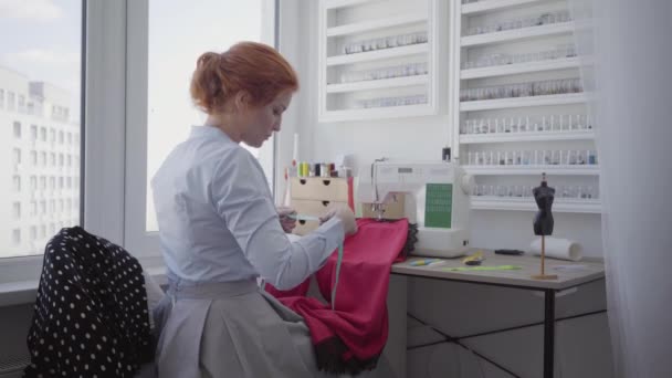 Beautiful red-haired woman sews clothes sitting at the table near the window in the light room. Seamstress works at home. Hobby. Sewing clothes. Shooting behind the curtain — Stock Video