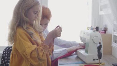 Küçük kızı ön planda bir fermuar ile oynarken onun kızıl saçlı annesi arka planda pencerenin yanındaki masada oturan giysileri diker. Terzi evde çalışıyor