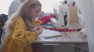 Küçük kız ön planda masada otururken kırmızı saçlı annesi arka planda yakın giysiler diker çizim. Terzi kızıyla evde çalışıyor. Mutlu aile birlikte