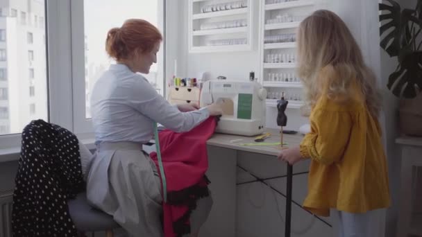 美しい赤毛の女性は、窓の近くのテーブルに座って服を縫い、明るい部屋で良い仕事をする方法を娘に示しています。シームレスは自宅で働いています。趣味。裁縫服 — ストック動画