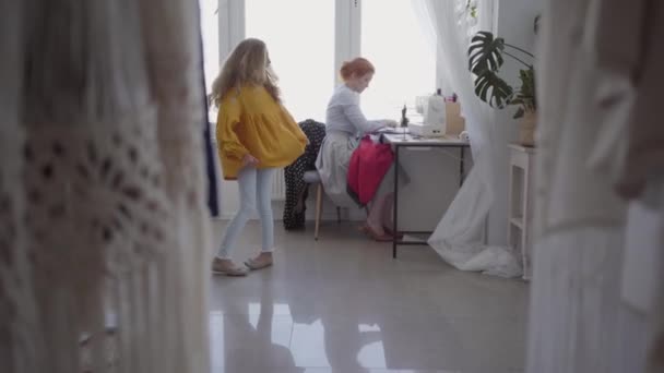 Gyönyörű vörös hajú nő SEWS ruhákat ül az asztalnál, közel az ablakon a fény szobába. Kislánya spinning körül közel. Varrónő dolgozik otthon. Hobbi. Varróruhák — Stock videók