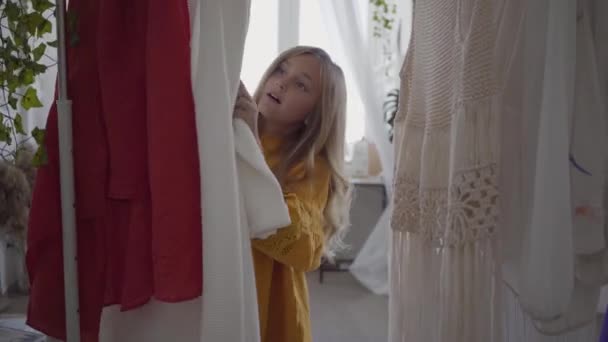 Menina bonito com cabelo longo escolher vestido em primeiro plano, enquanto sua mãe costura roupas sentadas no fundo. O Seamstress trabalha em casa. Hobby. Roupa de costura — Vídeo de Stock